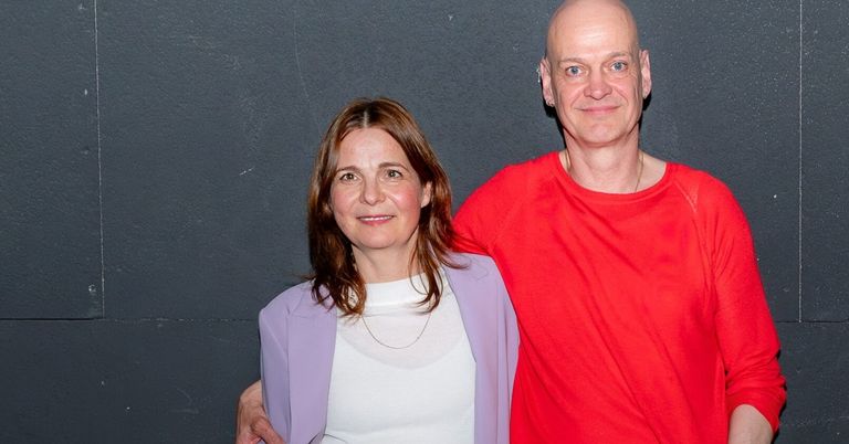 Ein Mann und eine Frau stehen nah beieinander und lächeln für die Kamera. Der Mann trägt ein rotes Shirt, und die Frau ein weißes Top und einen lila Blazer. Sie stehen vor einer grauen Wand.