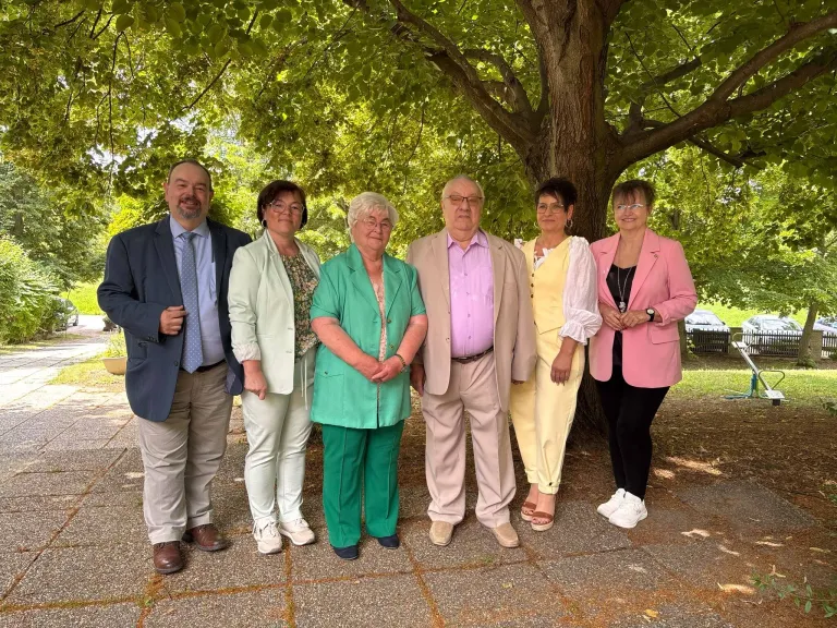 Sechs Erwachsene, zwei Männer und vier Frauen, stehen zusammen unter einem Baum. Sie sind in Sommerkleidung gekleidet. Sie lächeln für ein Foto.