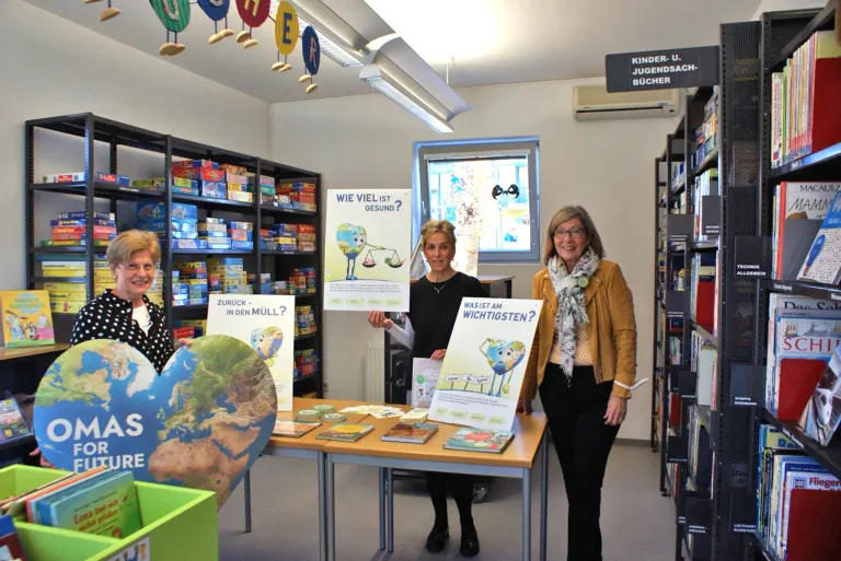 Drei Frauen stehen in einer Bibliothek, eine hält ein Plakat, das fragt: 'Wie viel ist gesund?' Ein anderes Plakat fragt: 'Was ist am wichtigsten?' Sie lächeln und stehen hinter einem Tisch mit Büchern.