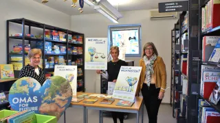 Drei Frauen stehen in einer Bibliothek, eine hält ein Plakat, das fragt: 'Wie viel ist gesund?' Ein anderes Plakat fragt: 'Was ist am wichtigsten?' Sie lächeln und stehen hinter einem Tisch mit Büchern.