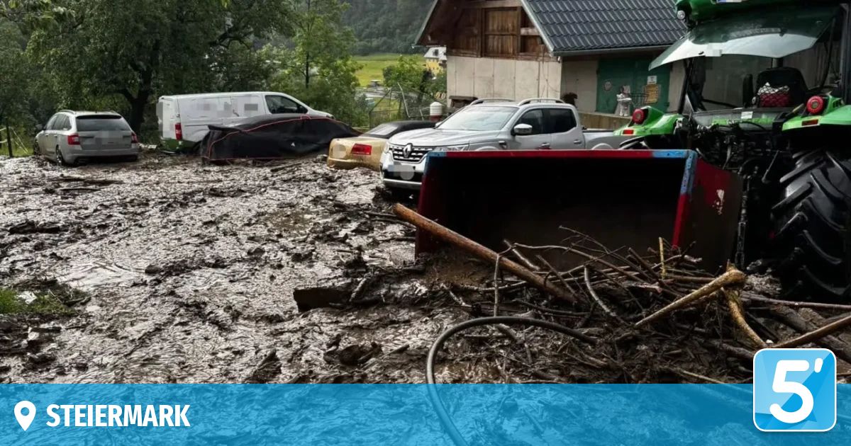 Schlamm und Schutt bedecken eine Straße mit mehreren Fahrzeugen. Ein Baufahrzeug reinigt den Bereich. Hinter den Fahrzeugen ist ein Haus mit grünem Dach zu sehen.