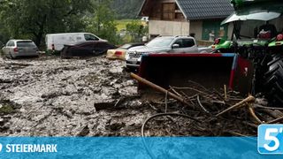 Schlamm und Schutt bedecken eine Straße mit mehreren Fahrzeugen. Ein Baufahrzeug reinigt den Bereich. Hinter den Fahrzeugen ist ein Haus mit grünem Dach zu sehen.