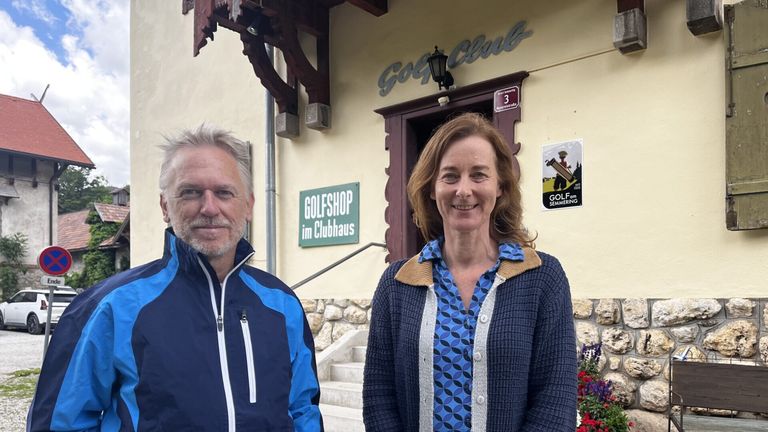 Zwei Personen stehen lächelnd vor einem Golfshop. Der Mann trägt eine blaue Jacke, und die Frau eine blaue Strickjacke. Dahinter befindet sich ein Gebäude mit einem grünen Schild 'Golfshop' und einem 'Golf Club'-Schild.