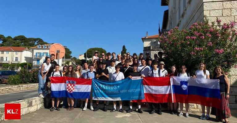 Eine Gruppe von Studenten hält Flaggen aus Kroatien und Österreich. Sie lächeln und stehen zusammen und halten ein blaues Banner mit der Aufschrift Erasmus+. Dahinter befindet sich ein Gebäude mit rosa Blumen.