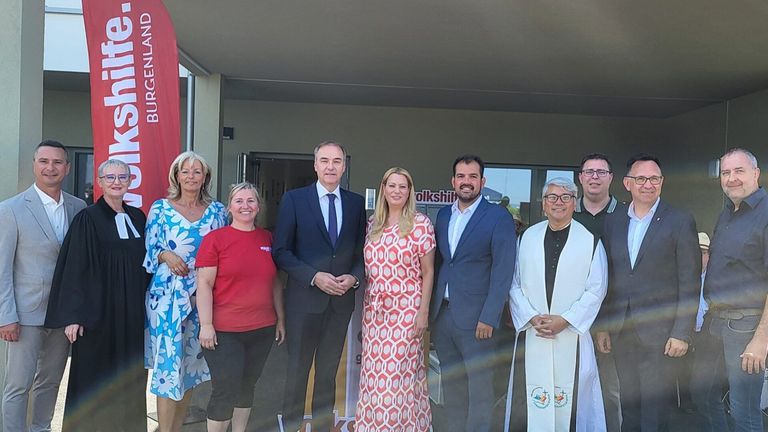 Eine Gruppe von Menschen posiert für ein Foto vor einem Gebäude. Sie tragen alle formelle Kleidung. Eine Person trägt ein rotes Shirt und zwei Personen tragen Anzüge und Krawatten. Eine weitere Person trägt eine weiße Robe.