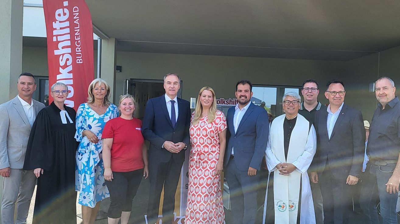 Eine Gruppe von Menschen posiert für ein Foto vor einem Gebäude. Sie tragen alle formelle Kleidung. Eine Person trägt ein rotes Shirt und zwei Personen tragen Anzüge und Krawatten. Eine weitere Person trägt eine weiße Robe.