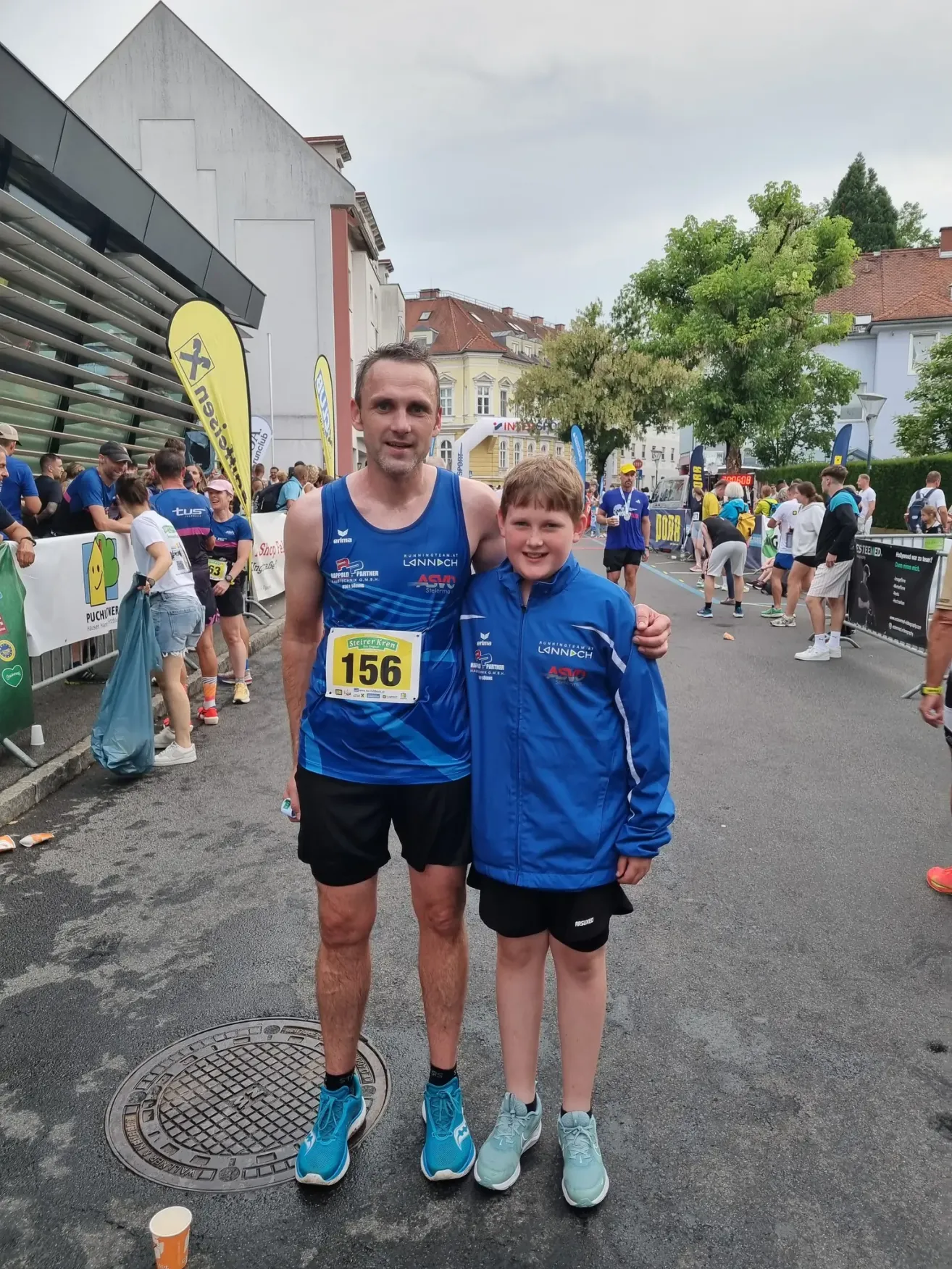 Ein Mann und ein Junge posieren für ein Foto bei einem Marathon-Event. Sie tragen passende blaue Jacken und Shorts. Gebäude, Bäume und Menschen säumen die Straße.