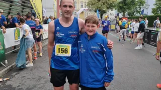Ein Mann und ein Junge posieren für ein Foto bei einem Marathon-Event. Sie tragen passende blaue Jacken und Shorts. Gebäude, Bäume und Menschen säumen die Straße.