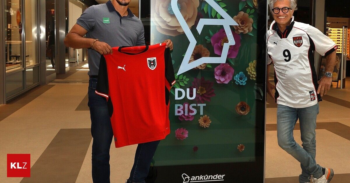 Zwei Männer stehen bei einem Plakat mit Blumendesign. Einer hält ein rotes Sporttrikot. Es steht 'DU BIST'. Der Mann links trägt ein graues Poloshirt. Der Mann rechts trägt ein weißes Trikot.