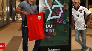 Zwei Männer stehen bei einem Plakat mit Blumendesign. Einer hält ein rotes Sporttrikot. Es steht 'DU BIST'. Der Mann links trägt ein graues Poloshirt. Der Mann rechts trägt ein weißes Trikot.