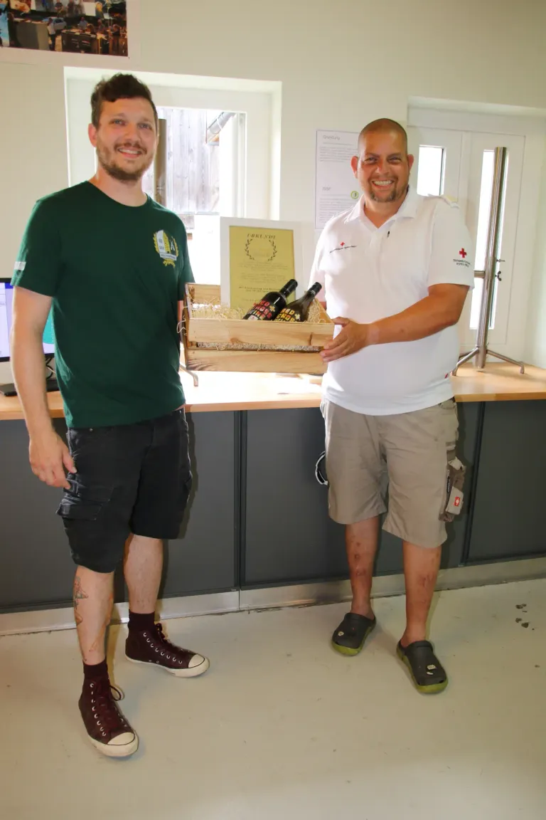 Two men, one in a green shirt, the other in a white polo shirt, hold a wooden box with wine bottles inside. They stand in front of a desk with a computer monitor.
