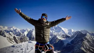 Ein Mann steht auf dem Gipfel einer Bergkette, Arme ausgestreckt, lächelt in die Kamera. Er ist in einer Winterjacke, Hose und Stiefeln gekleidet. Der Berg ist mit Schnee bedeckt.