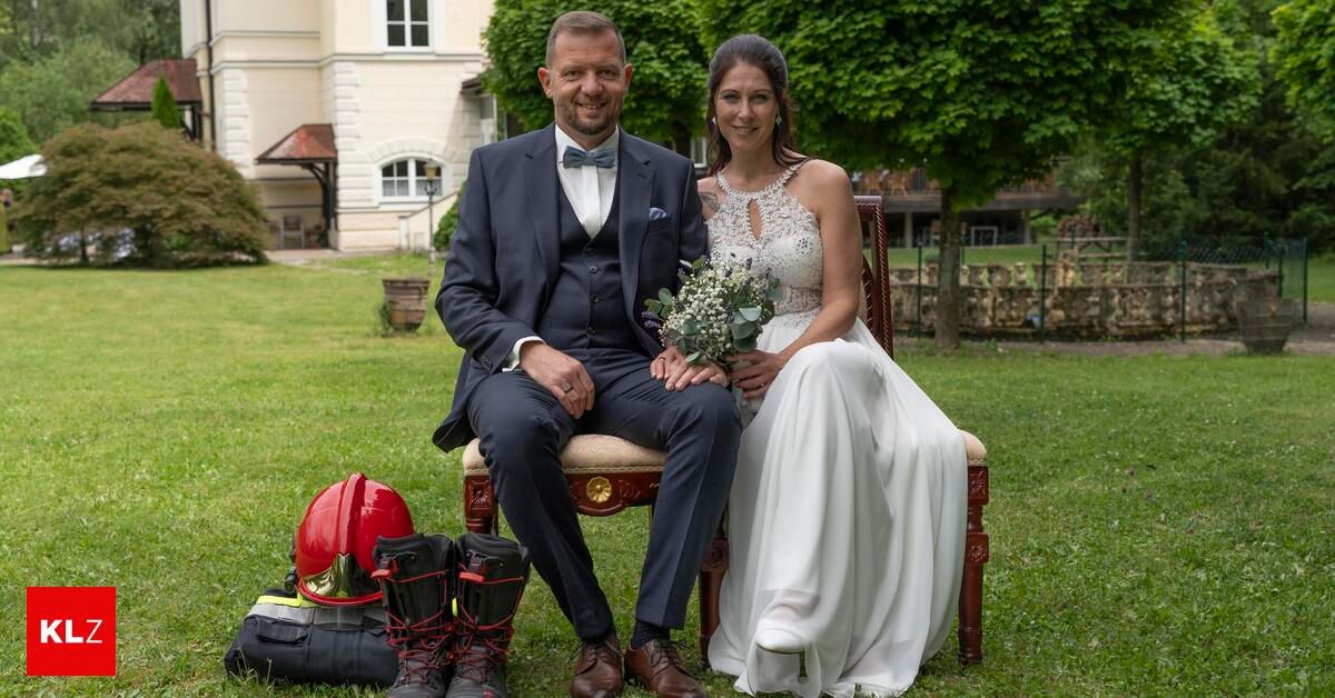 Ein Mann und eine Frau in formeller Kleidung sitzen auf einem Stuhl in einem Park. Sie halten Händchen. Die Frau hält einen Blumenstrauß in den Händen.
