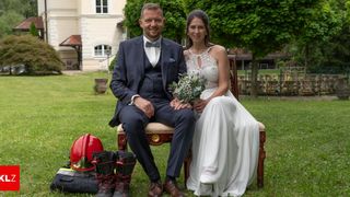 Ein Mann und eine Frau in formeller Kleidung sitzen auf einem Stuhl in einem Park. Sie halten Händchen. Die Frau hält einen Blumenstrauß in den Händen.