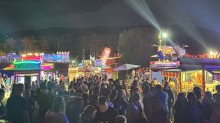 Eine Menschenmenge versammelt sich auf einem Nachtfestival, beleuchtet von bunten Lichtern. Fahrten und Essensbuden sind sichtbar, mit Bäumen und Bergen im Hintergrund.