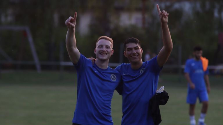 Zwei junge Männer in blauen Fußballtrikots posieren für ein Foto, lächeln und zeigen mit ihren Fingern.