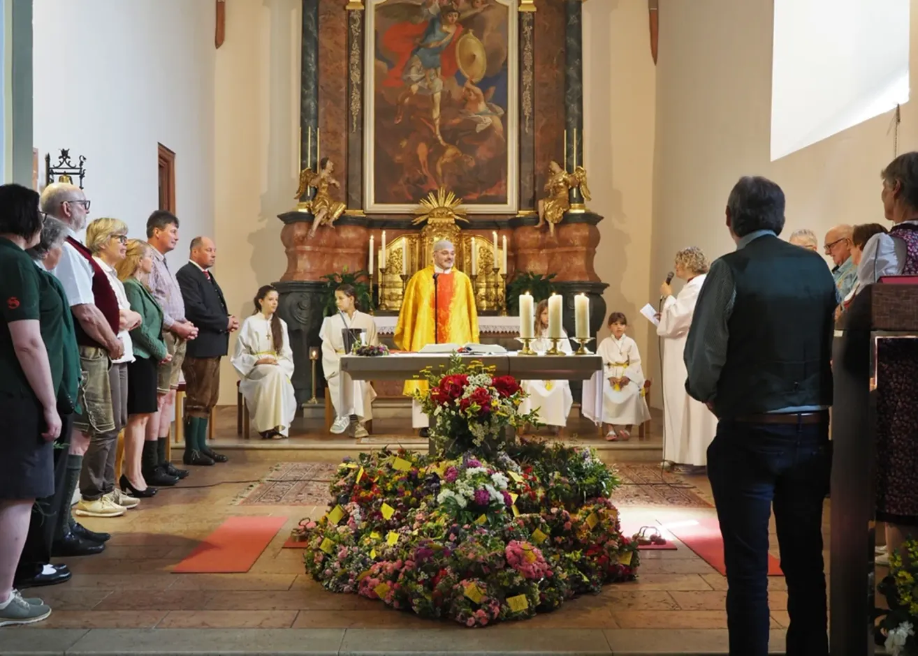 Ein Priester steht in einer Kirche am Altar mit einer Menschenmenge, Kerzen und Blumen. Ein großes Gemälde ist hinter ihm.