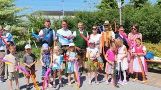 Eine Gruppe von Erwachsenen und Kindern steht zusammen und hält bunte Bänder in der Hand, vor einem Hintergrund aus Grünpflanzen und einem Gebäude. Sie lächeln und posieren für ein Foto.