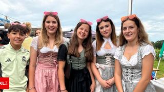 Fünf Frauen in traditioneller deutscher Kleidung, lächeln für die Kamera. Eine trägt ein rosa Kleid, eine andere ein schwarzes Kleid mit einem karierten Oberteil. Beide haben rosa Sonnenbrillen auf den Köpfen.
