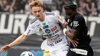 Zwei Fußballspieler in Aktion; einer in Weiß mit Werbeaufklebern, der andere in Schwarz, kämpfen auf dem Feld. Zuschauer im Hintergrund.