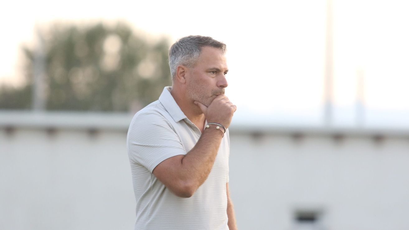 Ein Mann in einem weißen Hemd mit einem Armband am linken Handgelenk steht und denkt. Er berührt sein Kinn mit der linken Hand.