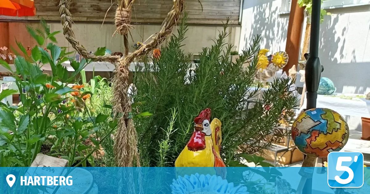 Ein Garten mit einem herzförmigen Strohkranz, einer bunten Hahnenstatue und verschiedenen Pflanzen und Blumen.