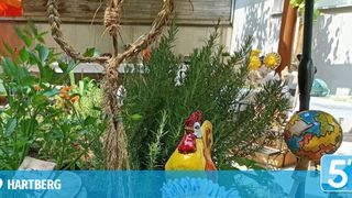 Ein Garten mit einem herzförmigen Strohkranz, einer bunten Hahnenstatue und verschiedenen Pflanzen und Blumen.