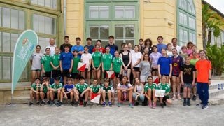 Eine Gruppe junger Menschen in verschiedenen farbigen Trikots und Turnschuhen posiert für ein Foto vor einem gelben Gebäude mit grünen Fenstern.