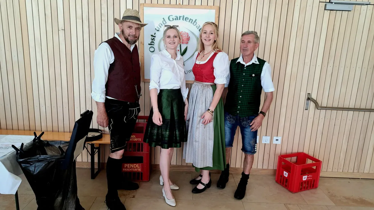 Vier Personen stehen zusammen, gekleidet in traditioneller Kleidung. Zwei Männer tragen Westen und Hüte, während zwei Frauen Kleider tragen. Hinter ihnen ist eine Holzwand mit einem Schild und einer Obstkiste.