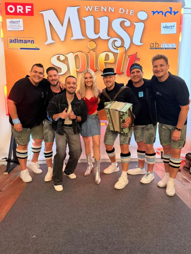 Sechs Erwachsene posieren für ein Foto vor einem Banner, das 'Mast spielt' steht. Vier Männer und zwei Frauen, einer der Männer hält ein Akkordeon. Sie tragen alle Shorts mit kniehohen Socken.
