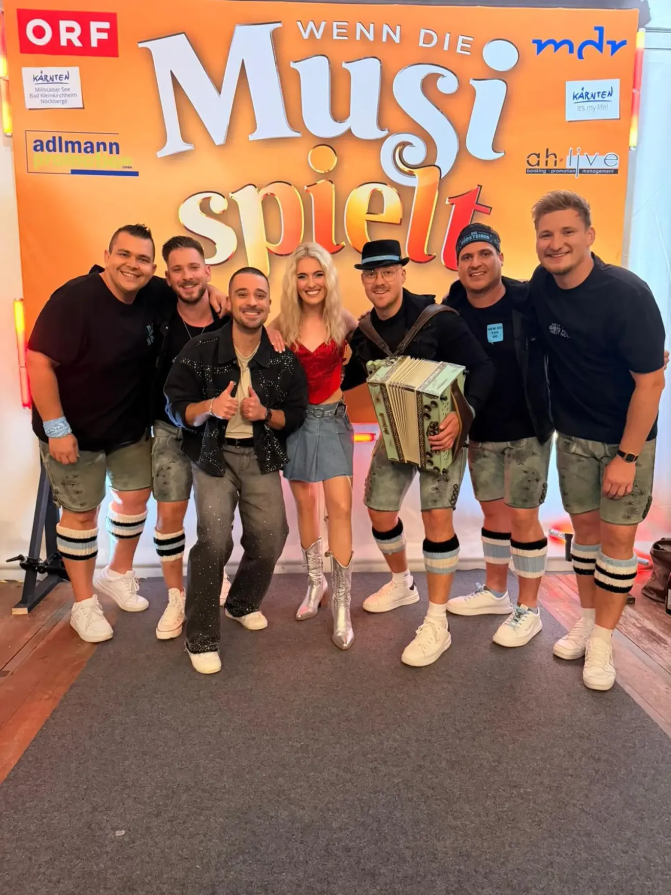 Sechs Erwachsene posieren für ein Foto vor einem Banner, das 'Mast spielt' steht. Vier Männer und zwei Frauen, einer der Männer hält ein Akkordeon. Sie tragen alle Shorts mit kniehohen Socken.