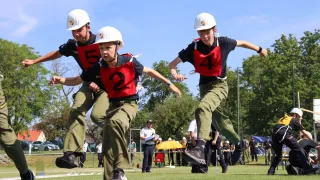 Drei junge Menschen mit Helmen und nummerierten Westen nehmen an einer Sportveranstaltung auf einem Feld teil. Im Hintergrund stehen Menschen, einer hält einen Regenschirm.