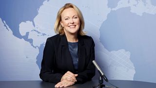 Eine Frau mit blonden Haaren sitzt an einem Schreibtisch, trägt einen schwarzen Blazer und lächelt. Sie steht vor einem Mikrofon, mit einer Weltkarte im Hintergrund.