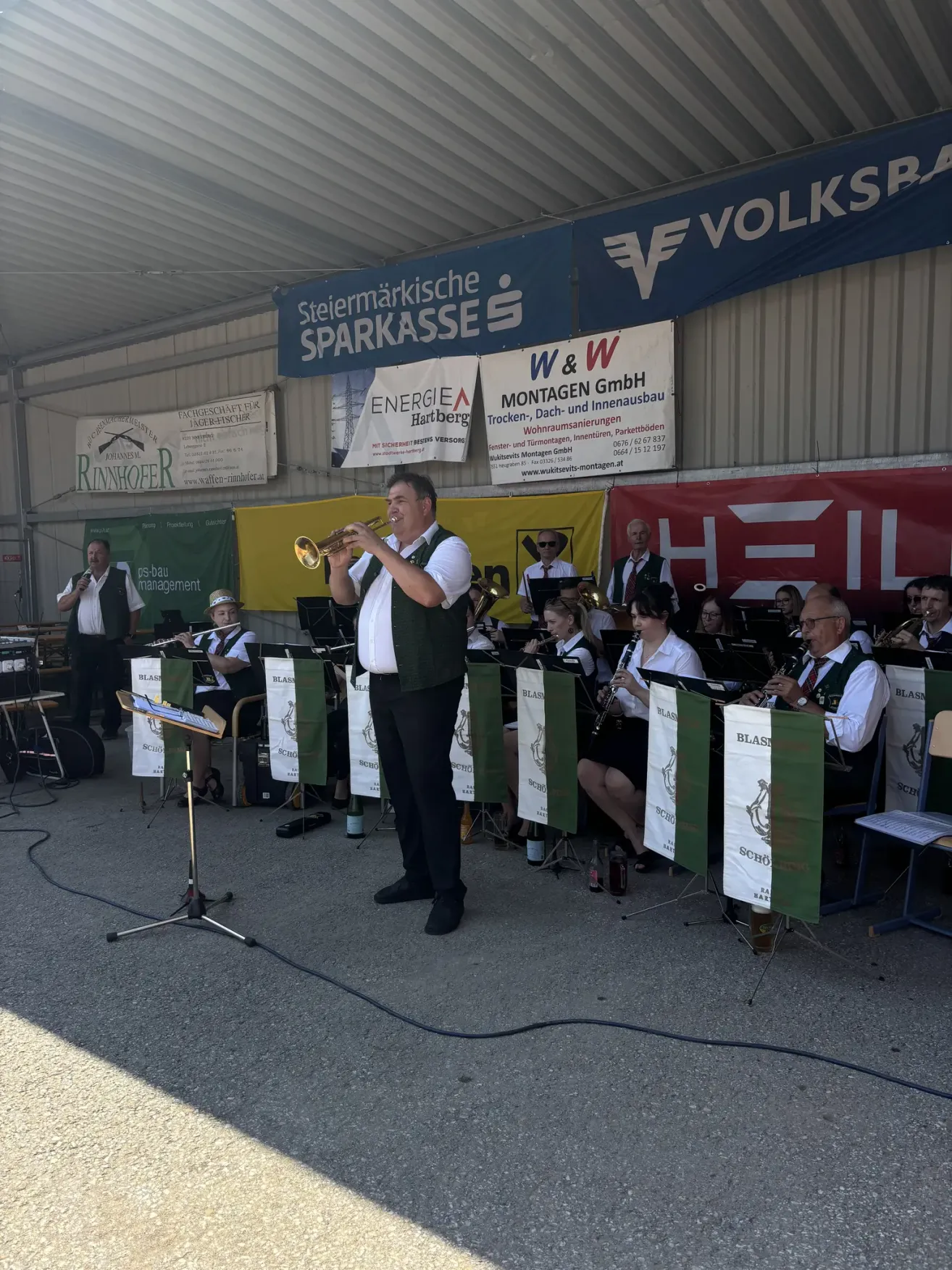 Eine Blaskapelle tritt auf der Bühne auf, ein Schild mit 'Steiermarkische Sparkasse'. Ein Mann steht vorne und spielt Trompete, während andere Instrumente hinter ihm spielen. Mehrere Zuschauer sind vor der Band sitzend.