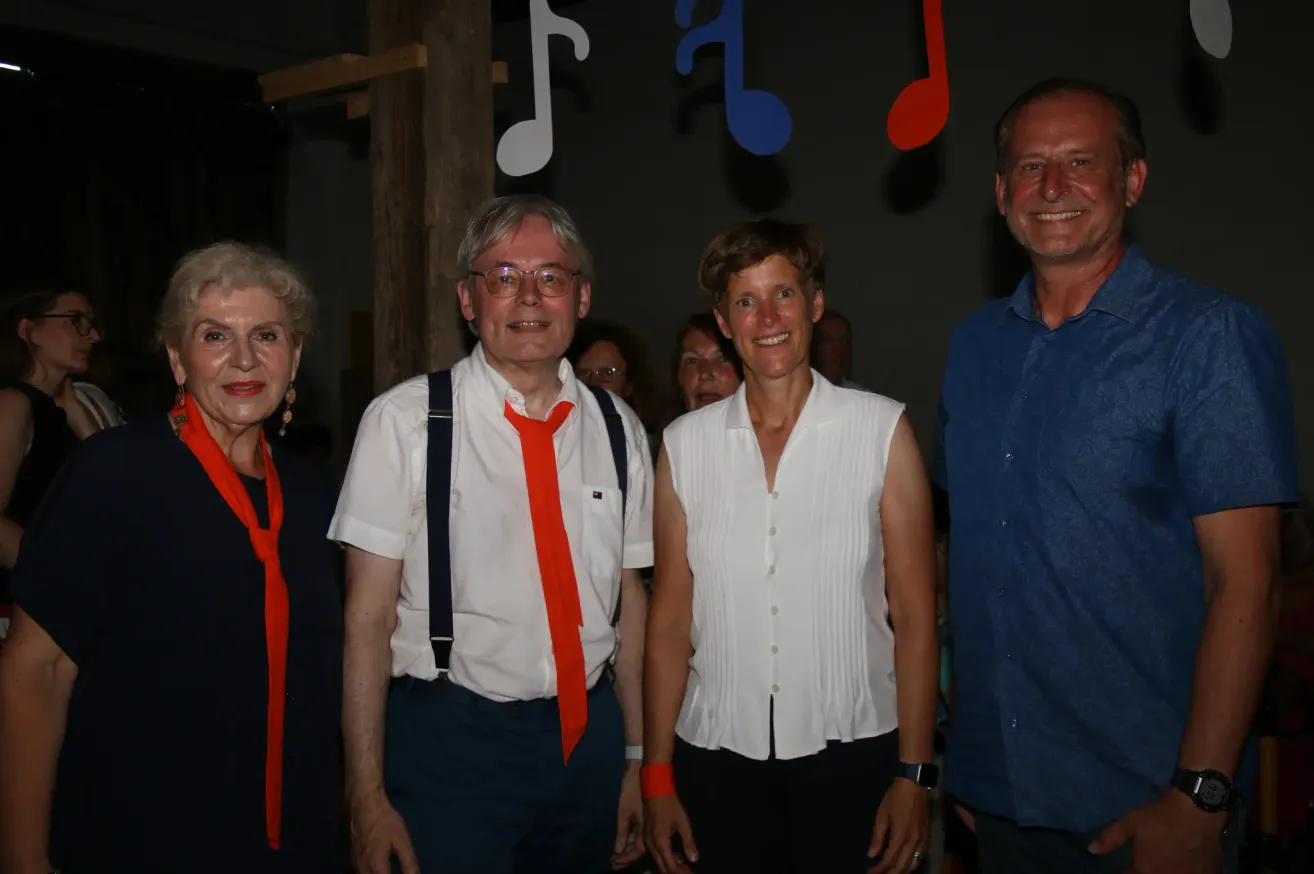 Vier Erwachsene, zwei Männer und zwei Frauen, stehen eng beieinander für ein Foto in einer festlichen Umgebung mit musikalischen Noten im Hintergrund.