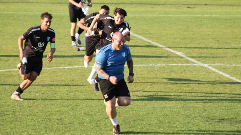 Ein Mann in Blau rennt auf einem Fußballfeld, verfolgt von einem anderen Spieler in Schwarz, während ein dritter Spieler in Weiß eine Flasche hält.