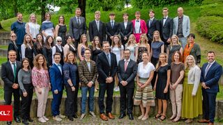 Eine große Gruppe formell gekleideter Männer und Frauen, wahrscheinlich Fakultätsmitglieder und Studenten, posieren für ein Foto an einem sonnigen Tag vor einer Steinmauer mit Grünpflanzen im Hintergrund.