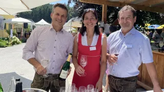 Drei Personen stehen bei einer Outdoor-Veranstaltung mit einem Tisch und Weinläsern davor. Ein Mann auf der linken Seite hält eine Flasche Wein. Eine Frau in einem roten Kleid steht in der Mitte und hält ein Weinglas. Ein Mann auf der rechten Seite hält ein Weinglas.
