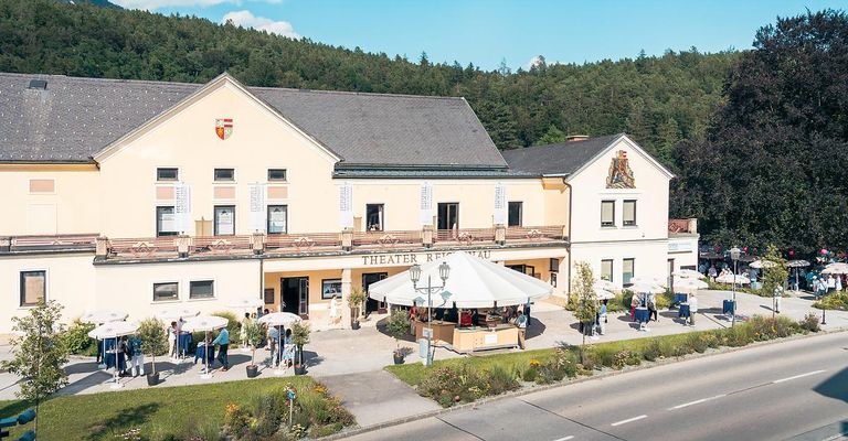 Luftaufnahme eines Theatergebäudes mit einem großen Zelt und Leuten draußen. Das Gebäude hat ein Wappen an seiner Fassade. Bäume und ein Berg sind im Hintergrund.
