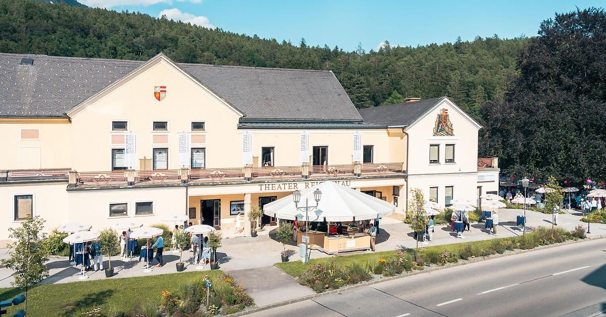 Luftaufnahme eines Theatergebäudes mit einem großen Zelt und Leuten draußen. Das Gebäude hat ein Wappen an seiner Fassade. Bäume und ein Berg sind im Hintergrund.