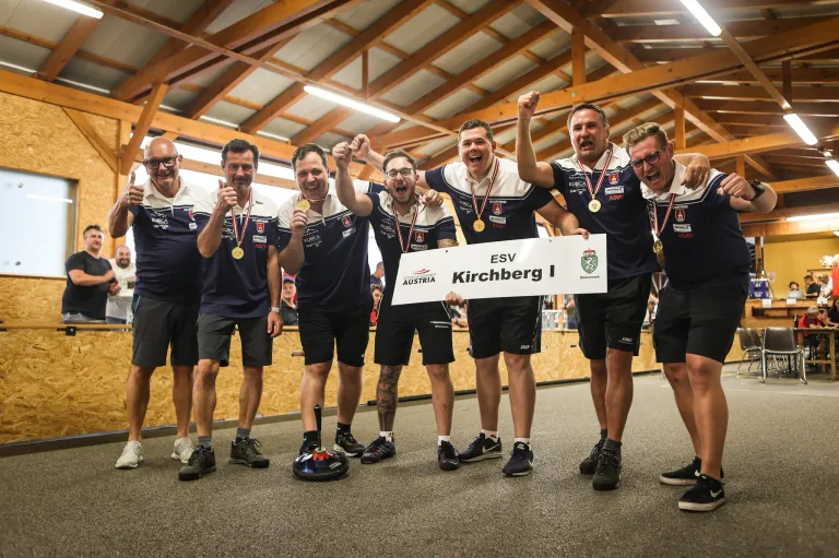 Sieben Männer feiern mit Goldmedaillen, stehen in einer Reihe, halten ein Schild mit ESV Kirchberg I. Sie tragen passende Uniformen und sind in einem Raum mit Holzbalken.
