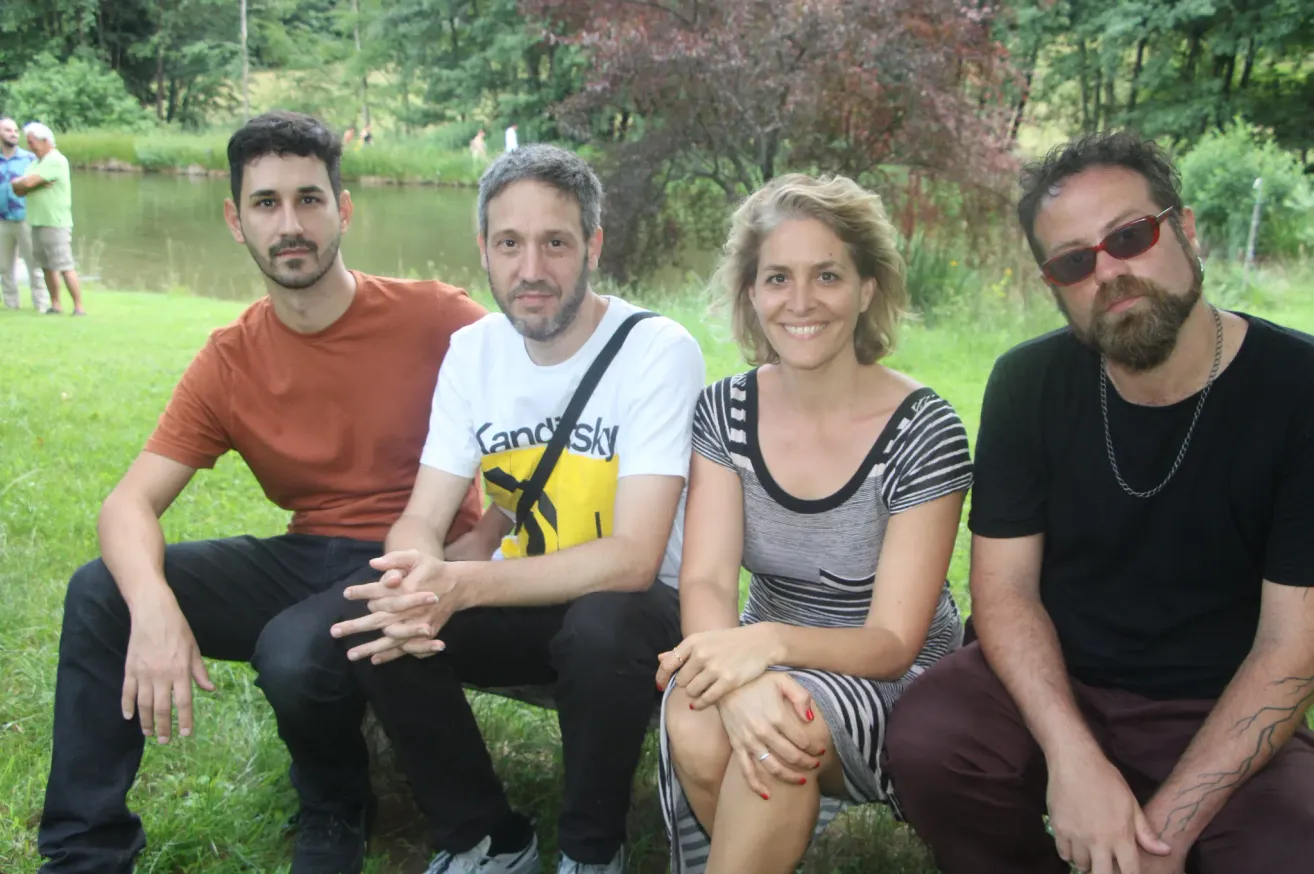 Vier Menschen sitzen lächelnd auf dem Rasen. Ein Mann mit Rucksack lächelt, eine Frau trägt ein gestreiftes Kleid und ein Mann mit Sonnenbrille. Im Hintergrund ist ein Teich und Bäume zu sehen.