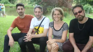 Vier Menschen sitzen lächelnd auf dem Rasen. Ein Mann mit Rucksack lächelt, eine Frau trägt ein gestreiftes Kleid und ein Mann mit Sonnenbrille. Im Hintergrund ist ein Teich und Bäume zu sehen.