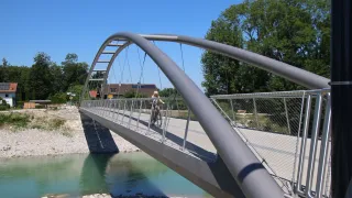 Ein Radfahrer fährt auf einem kurvigen Metallbogen über einen Fluss, umgeben von üppigem Baumbestand und einem klaren Himmel.