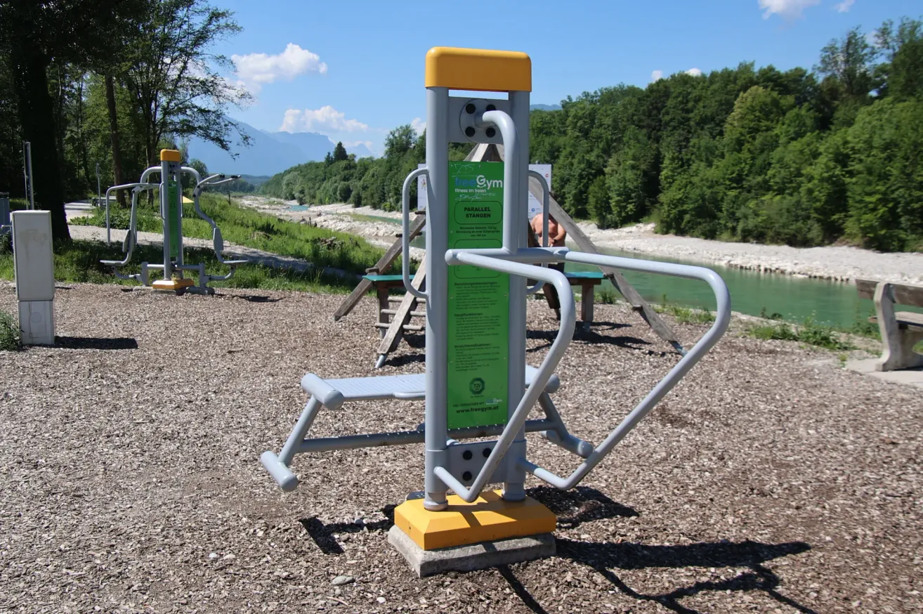 Eine Outdoor-Trainingsstation mit grünen Anweisungen und einer gelben Basis. Dahinter ein Fluss und ein Grasbereich mit Bäumen und Bergen.