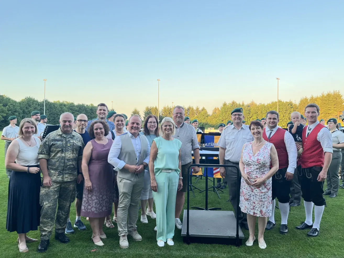 Eine Gruppe von Menschen steht auf einem Grasfeld und posiert für ein Foto. Sie tragen verschiedene Outfits, und einige tragen Hüte. Sie stehen in der Nähe eines Notenständers, und einige tragen Militäruniformen. Im Hintergrund befinden sich Straßenlaternen und Bäume.
