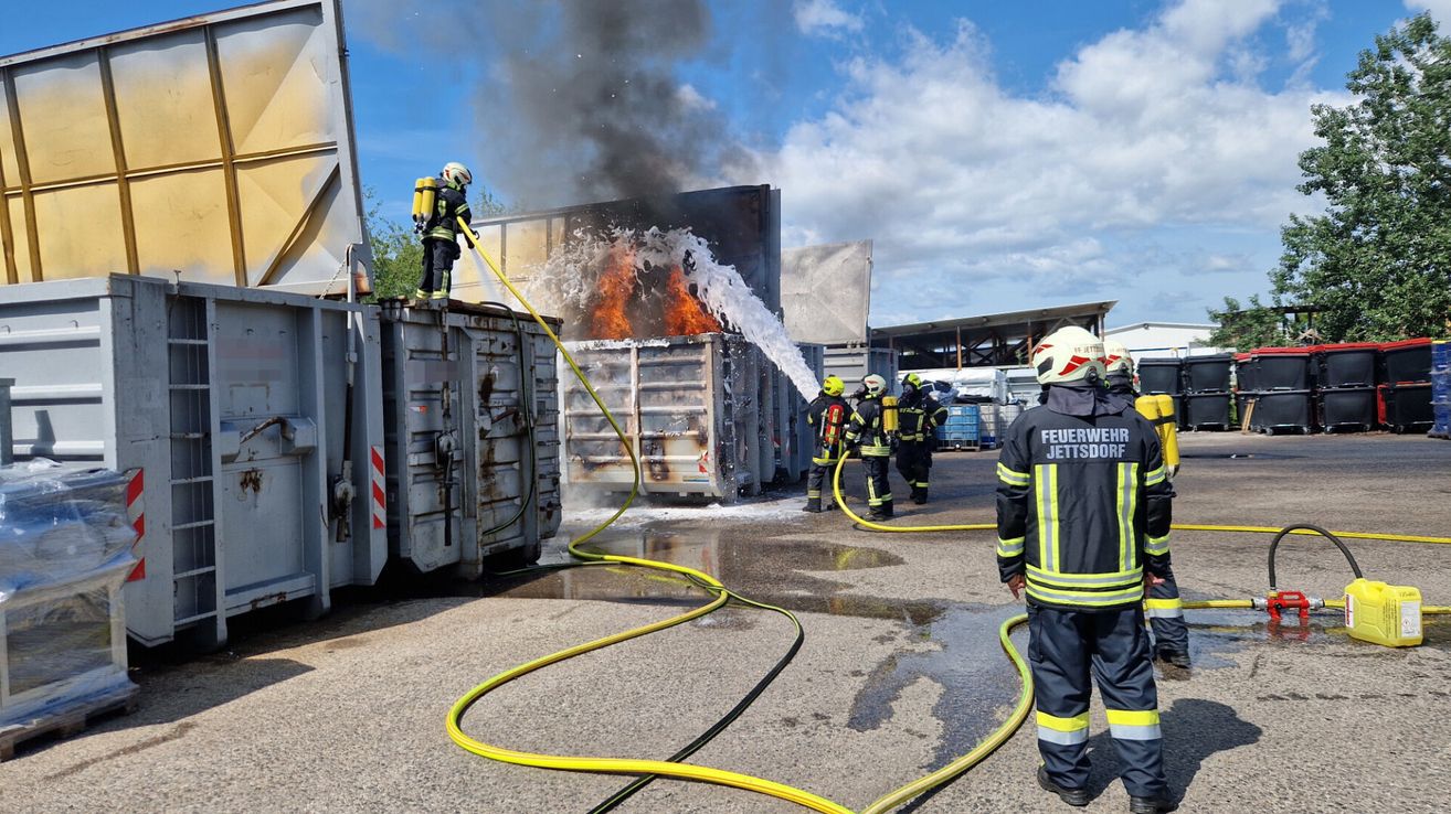 Feuerwehrleute spritzen mit einem Schlauch Wasser auf einen brennenden Müllcontainer, wobei Flammen und Rauch sichtbar sind.
