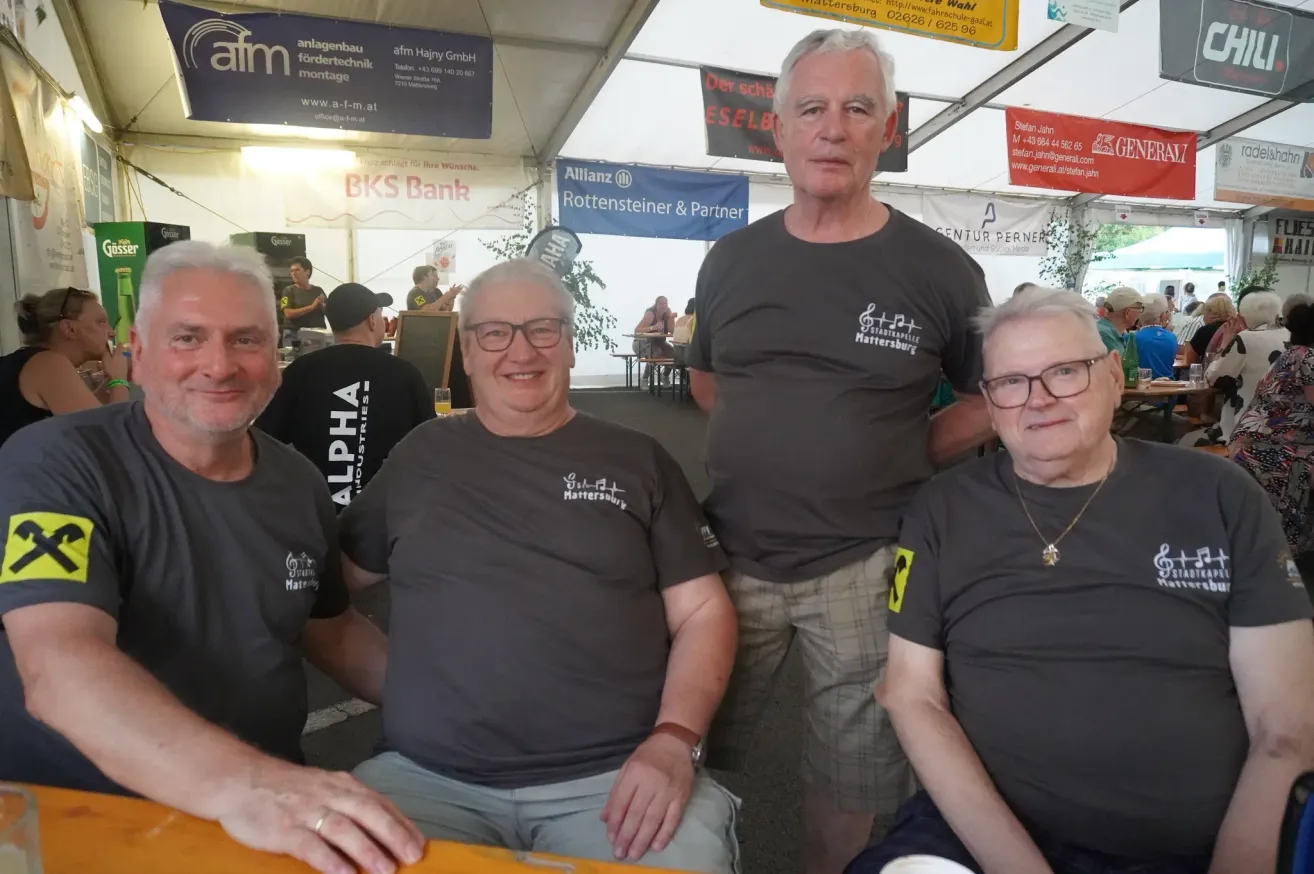 Vier Männer sitzen unter einem Zelt. Der Mann in der Mitte trägt ein graues T-Shirt mit weißen Text. Es gibt Schilder am Zelt. Dahinter sitzt eine Person an einem Tisch.