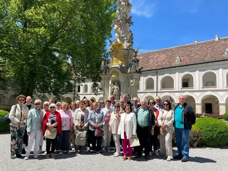 Eine Gruppe von Erwachsenen steht vor einem kunstvollen Brunnen und posiert für ein Foto. Sie tragen Freizeitkleidung und einige haben Sonnenbrillen auf. Hinter ihnen befindet sich ein Gebäude mit Bogenfenstern und einer Statue oben.