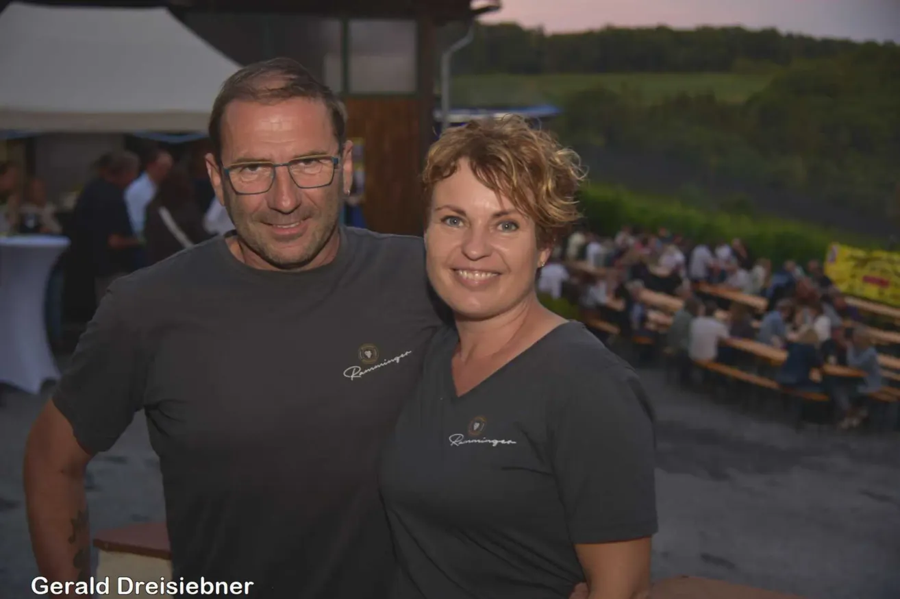 Ein Mann und eine Frau, beide in schwarzen T-Shirts mit einem Logo, stehen eng zusammen und lächeln bei einer Outdoor-Veranstaltung. Im Hintergrund sitzen viele Menschen an Tischen.